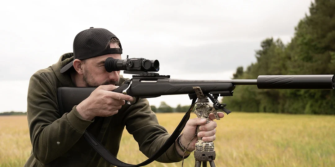 Hunter aiming a bolt-action rifle mounted with a DNT Optics ZHD520R-V2 scope, prone with a bipod in a grain field